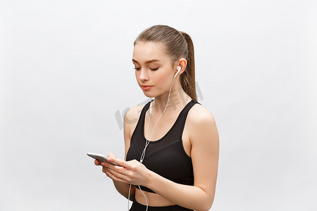 年轻女运动员的孤立镜头拥有健康的运动身体，用耳机听音乐，拿着智能手机。