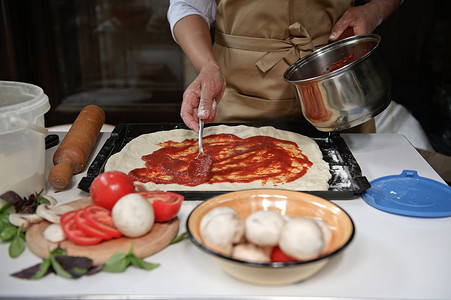 身穿米色厨师围裙的前视家庭主妇将新鲜制作的番茄酱涂在擀好的面团上。
