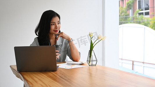 坐着的会计摄影照片_体贴的女商人坐在笔记本电脑前，望着窗外，梦想着未来新的成功项目