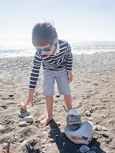 小男孩海边摄影照片_小男孩正在圆石滩上用石头和浮木建造金字塔。