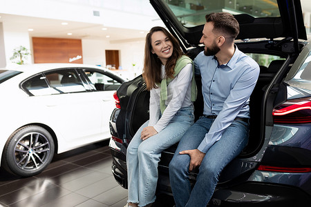 汽车经销店里的一对年轻夫妇选择了一辆微笑着坐在跨界车后备箱里的汽车