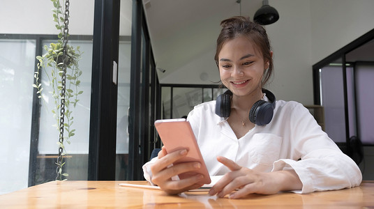 挥舞江湖摄影照片_快乐的亚洲年轻女孩坐在家里的沙发上，拿着手机，看着屏幕，挥舞着手，使用智能手机视频聊天应用程序，在移动聊天应用程序中在线与远方的朋友进行视频通话。