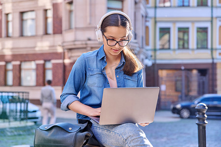 戴耳机的少女学生在户外、城市背景下使用笔记本电脑