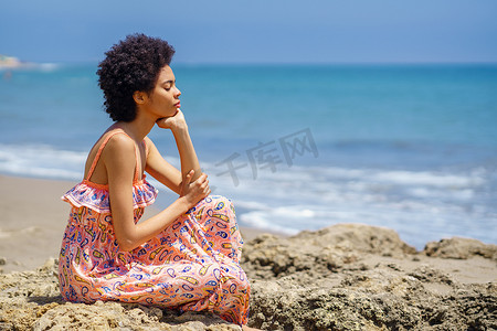 年轻的黑人女子坐在海滩上的岩石上，闭着眼睛，用正念技巧放松。