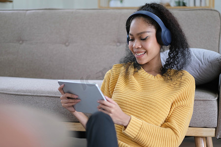 照片中，美丽的深色皮肤波浪女士温馨心情拿着数字平板电脑耳罩，坐在客厅沙发休闲服黄色附近的地板上听新音频