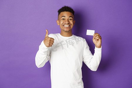 银行贷款摄影照片_惊讶的非洲裔美国人竖起大拇指和信用卡的肖像，推荐银行贷款，站在紫色背景下
