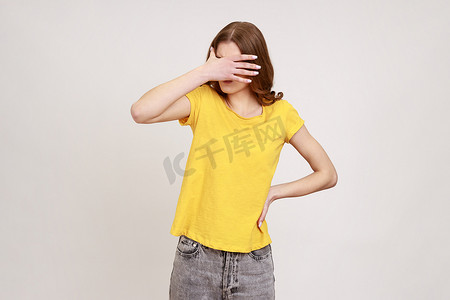身穿休闲黄色 T 恤的年轻棕色头发女子站着，用手闭上眼睛，不想看到任何东西。