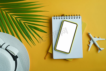 黄色背景上的智能手机、帽子、笔记本和绿叶热带棕榈。