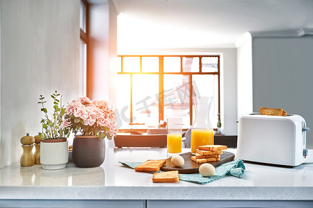 面包鸡蛋橙汁摄影照片_烤面包机配有新鲜面包、鸡蛋和一杯橙汁，放在浅色的厨房桌子上。