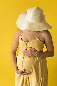 复古摩擦摄影照片_母性、女性气质、时尚、炎热的夏天 — 裁剪特写镜头无法辨认的孕妇女士穿着漂亮的复古连衣裙普罗旺斯草帽，双手放在肚子上，黄色背景上摩擦着肚子