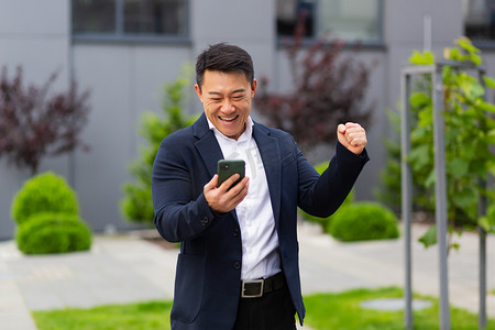 亚洲商人商人看着手机阅读彩票中奖消息在办公室外欢欣鼓舞