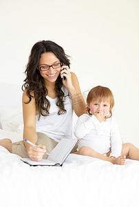 重要日程摄影照片_良好的日程安排对于单亲父母来说很重要......一位年轻的母亲在手机上说话，在笔记本上写字，而她的孩子和她一起坐在床上 - 肖像。