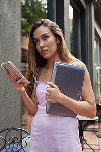手机夏季夏季摄影照片_穿着粉色裙子的年轻亚洲女性拿着笔记本电脑站在户外，使用智能手机