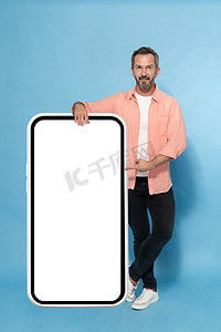 中年灰发男子靠在巨大的智能手机上，指着白色屏幕，穿着蓝色背景的桃色衬衫，在镜头前开心地微笑着。