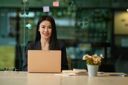 自信的女企业家坐在现代办公室里，配有笔记本电脑，对着镜头微笑