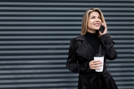 灰色星型摄影照片_一位穿着商务时尚服装的女商人的肖像，她拿着一杯咖啡，在灰色浮雕墙上用手机说话，这是商业成功和职业发展的概念