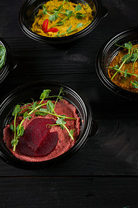 鹰嘴豆泥在深色木桌上的黑碗里装饰着辣椒、辣椒、甜菜和香草。