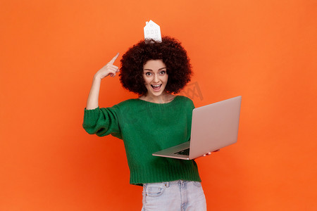 有趣而兴奋的女人，留着非洲发型，穿着绿色休闲风格的毛衣，指着头上的纸房子，用笔记本电脑。