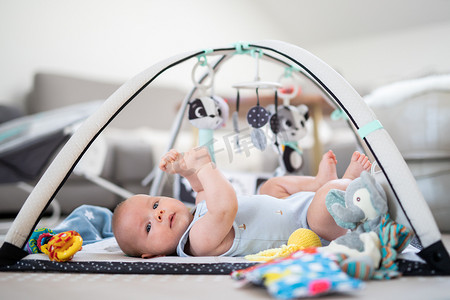 拱形贴标摄影照片_可爱的小男孩在家里的垫子上玩悬挂的玩具拱门 婴儿活动和游戏中心，用于婴儿早期发育。