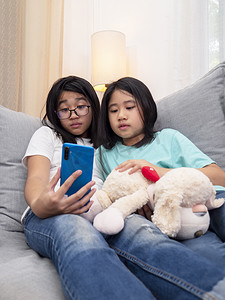 坐在客厅沙发上的快乐兄弟姐妹孩子们一起通过手机与父母交谈，微笑的大女孩通过智能手机展示可爱的小妹妹视频通话与家人交谈。
