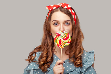 惊讶的海报女孩荷叶边上衣舔着甜甜的糖果，看着镜头，吃着美味的糖果棒棒糖，表情惊讶。
