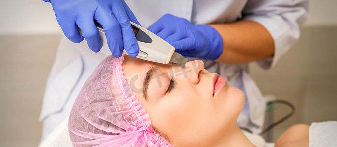 年轻的白人妇女在医疗沙龙接受超声波美容面部设备的面部皮肤清洁。