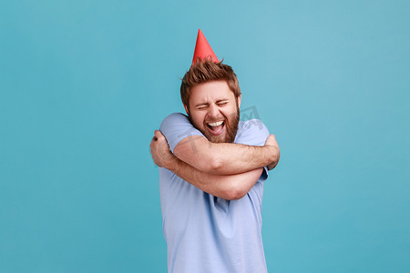 派对锥体中的男人用兴奋的面部表情拥抱自己，庆祝他的生日。