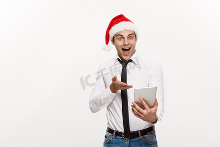 圣诞节概念-英俊的商人戴圣诞老人的帽子玩平板电脑与令人惊讶的面部表情。
