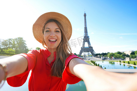 在法国巴黎，穿着红色裙子、戴着帽子的兴奋的时尚女性很高兴以埃菲尔铁塔为背景制作自画像。