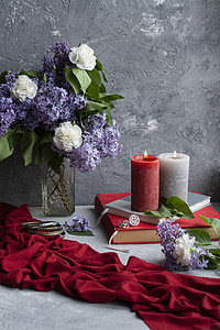 红色大气背景摄影照片_大气照片：灰色背景上的一束丁香和康乃馨、几根大蜡烛、书籍和珠宝