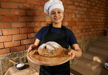 可爱的女人，戴着白色厨师帽和黑色围裙的面包师，递给镜头一块木板，上面放着全麦酵母面包
