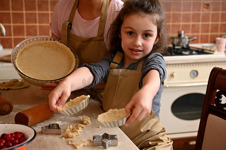 糖果展示摄影照片_妈妈和女儿穿着围裙，一起做饭，一边制作自制的馅饼和馅饼，一边在模具中展示卷好的面团