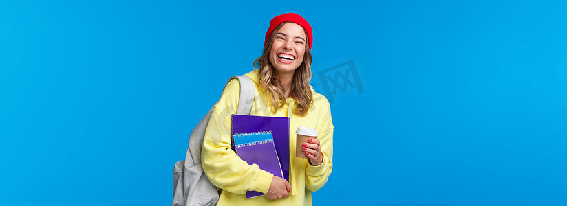 快乐快乐的女学生玩得开心，享受大学生活方式，背着背包和笔记本，喝外卖咖啡，与同学交谈，站立蓝色背景快乐