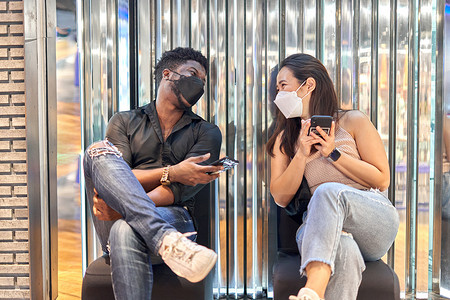 多民族朋友戴着面具，使用手机坐在购物中心