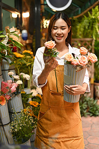 穿着围裙、手持粉红玫瑰花束站在花店前的积极的年轻女小企业家