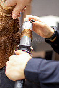 卷发女摄影照片_女发型师的手在美发沙龙里用卷发器给顾客卷发，特写。