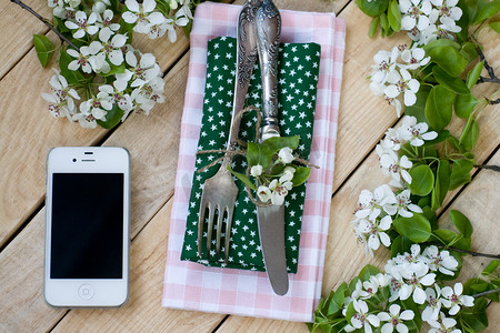 粉色苹果手机摄影照片_叉子和刀子，白色智能手机躺在木质背景上，周围环绕着鲜花。