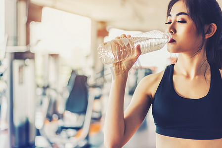 亚洲女性在有健身器材背景的健身房锻炼或运动训练后饮用纯净饮用水以保持新鲜。