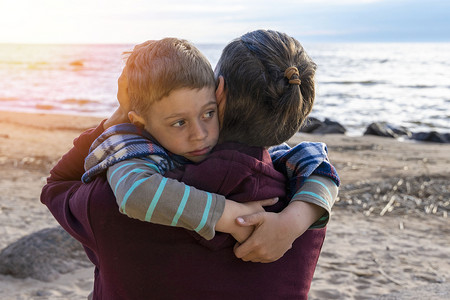 夕阳下的男孩摄影照片_可爱悲伤的学龄前男孩在夕阳下的海边拥抱他的爸爸