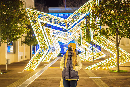 节日灯光摄影照片_晚上，女孩戴上针对冠状病毒COVID 19的医用口罩，在装饰有节日灯光的圣诞传统市场上散步。
