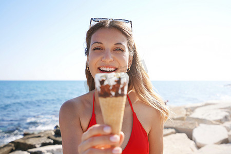 吃冰淇淋的女人摄影照片_夏天，美丽的比基尼女人拿着冰淇淋锥意大利冰淇淋的特写镜头，看着海滩上的相机