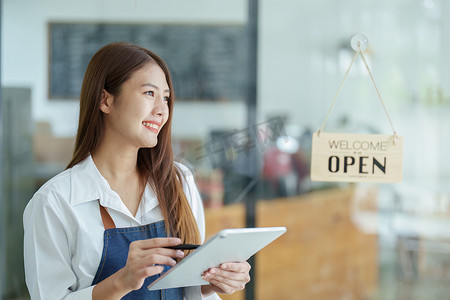一位美丽的亚洲女性经营一家小企业，手持平板电脑，脸上露出笑脸，一位咖啡店老板在新的早晨开店欢迎顾客，中小企业概念