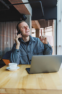 白天，身着休闲装的微笑男性企业家坐在桌子旁，拿着笔记本电脑，在带大窗户的现代工作空间中用智能手机交谈