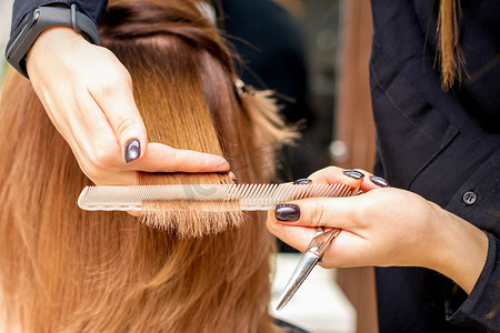 美发师的手将发束夹在手指之间，在美发沙龙里用梳子和剪刀为年轻女子的长发进行理发，特写。