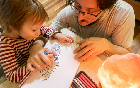 穿着灰色 T 恤的留着胡子的男人和可爱的男孩一起在家里度过时光，用彩色蜡笔画画。