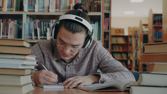 戴着大耳机的年轻白人男学生坐在图书馆的桌子旁。