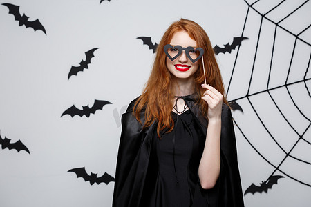 万圣节概念-穿着黑色女巫礼服的漂亮女孩拿着派对道具。