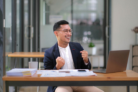 一位亚洲男性企业主站在电脑前的肖像，在成功投资后表现出幸福感。
