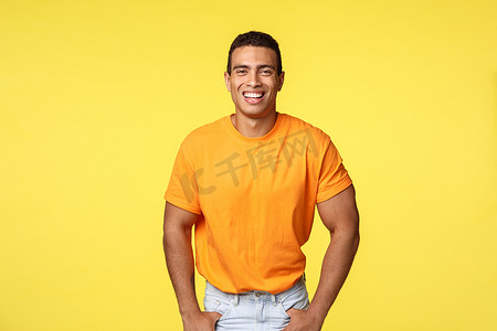 英俊开朗的微笑，穿着橙色T恤的阳刚男子快乐地笑着，微笑着友好的积极表情，手插在口袋里，随意地站在新年派对上，黄色背景
