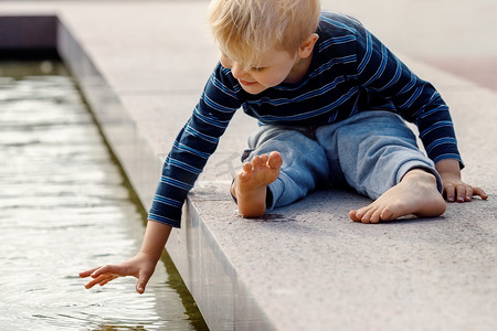 一个滑稽的小男孩爬上喷泉的护栏，触摸水。
